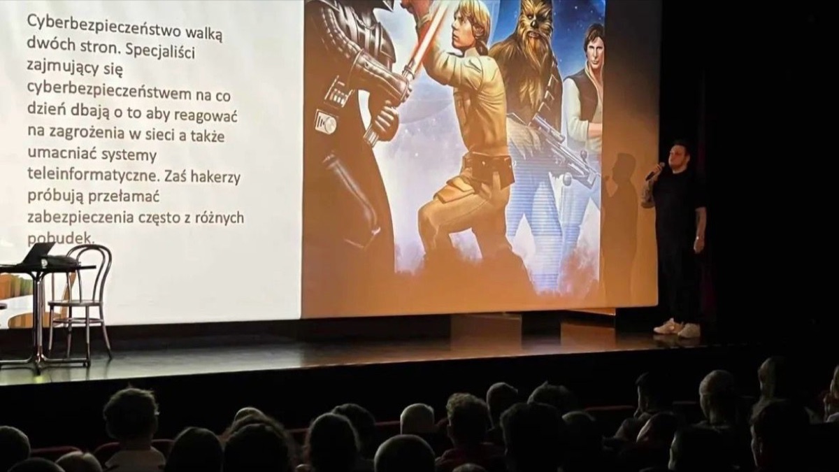 Jakub Lipiec na scenie przed salą pełną młodzieży, na ekranie slajd o cyberbezpieczeństwie